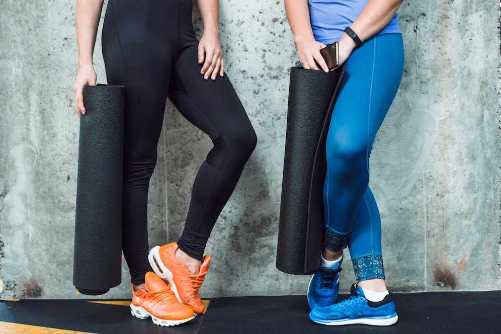 Two lower bodies of females wearing yoga pants, holding yoga mats.