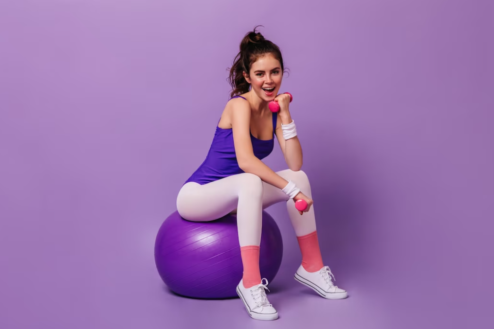 woman in pink leggings and blue body sitting on purple fitness ball in purple room